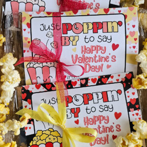 Popcorn Valentine Card tied onto microwave popcorn with a ribbon. Text on card reads, "Just Poppin' by to say Happy Valentine's Day." 3 valentines lined up with popcorn scattered around them.